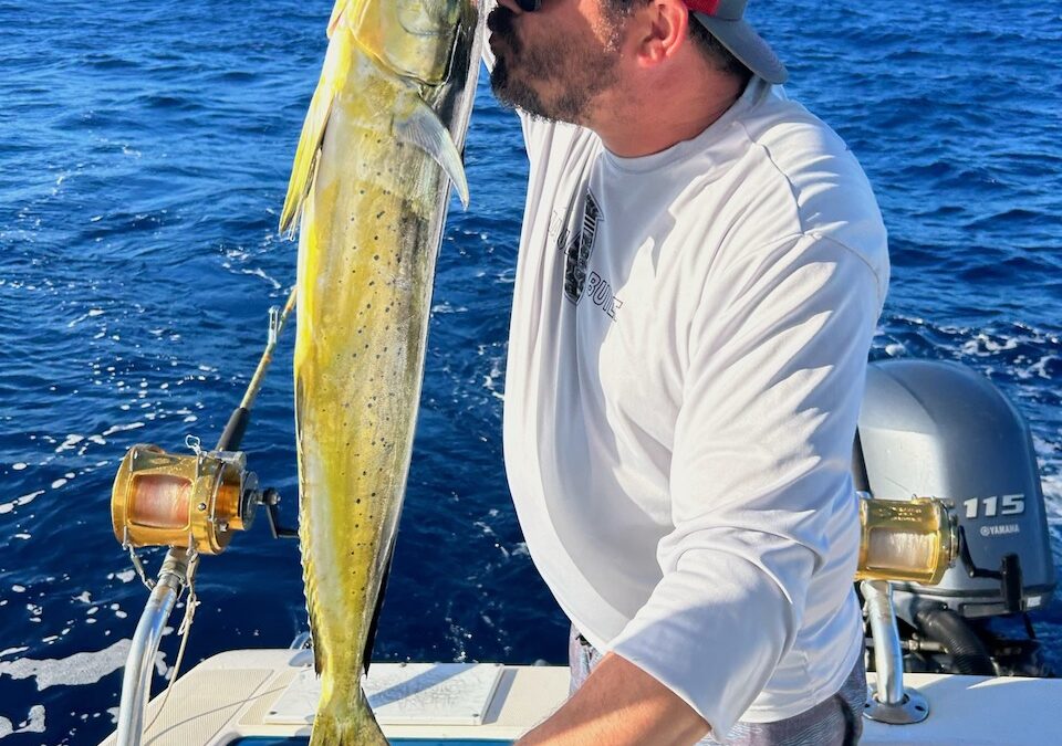 Alt Text: "A man kissing a mahi-mahi fish on a boat in the ocean, wearing a red cap and sunglasses."