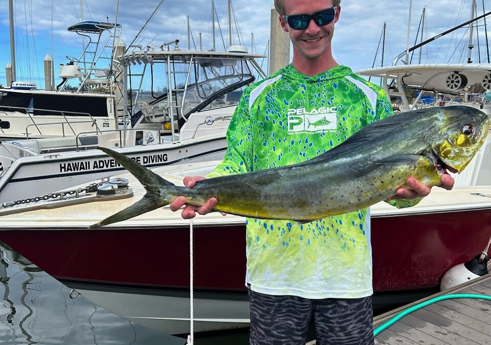 A young man in sunglasses and a tie-dye shirt proudly holds a large Mahi-mahi fish in front of a marina with boats docked in the background.